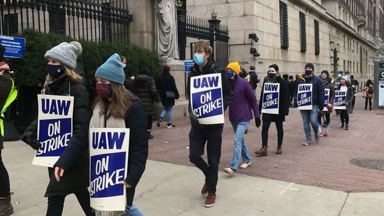 Columbia University postdocs and graduate workers picketing outside the campus gates on Broadway, carrying blue and white “UAW On Strike” signs, with Pulitzer Hall behind them.