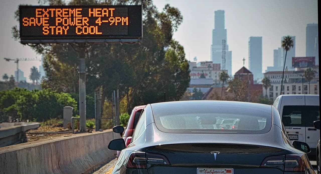 A digital highway sign reading ‘EXTREME HEAT, SAVE POWER 4-9PM, STAY COOL’ above a freeway, with the Los Angeles skyline visible in the smoggy background and cars in traffic below.