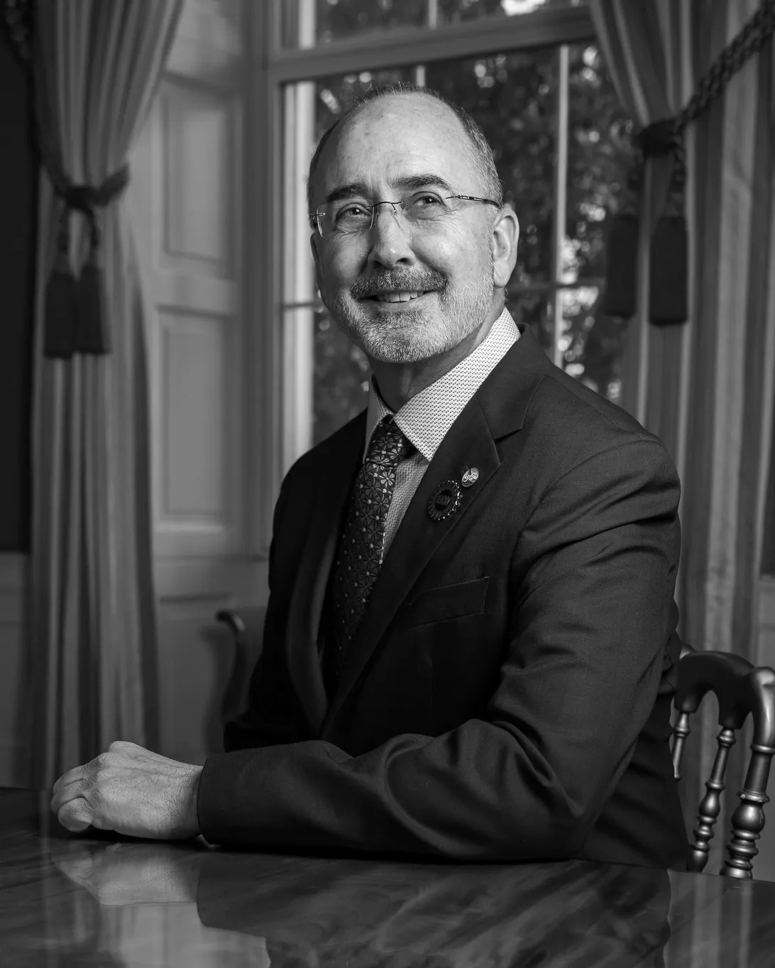 Black-and-white portrait of UAW President Shawn Fain, a balding man with glasses and a gray mustache and beard, seated at a table in a dark suit and patterned tie with UAW pins, in front of a curtained window.
