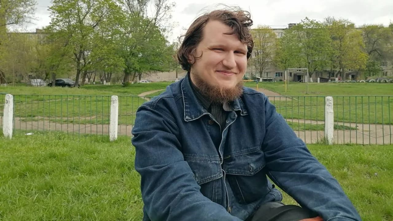 Bogdan Syrotiuk, a young man with wavy brown hair and a beard wearing a denim jacket, smiling while seated on grass in a park, with a green metal fence, trees, and a Soviet-era apartment building behind him.