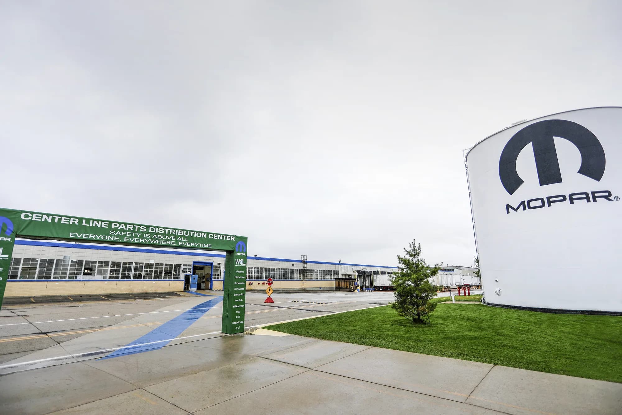 The entrance to Stellantis’ Mopar Center Line Parts Distribution Center under an overcast sky. A green banner over the walkway reads “CENTER LINE PARTS DISTRIBUTION CENTER — SAFETY IS ABOVE ALL. EVERYONE. EVERYWHERE. EVERYTIME,” and a large white silo alongside the building carries the Mopar logo.