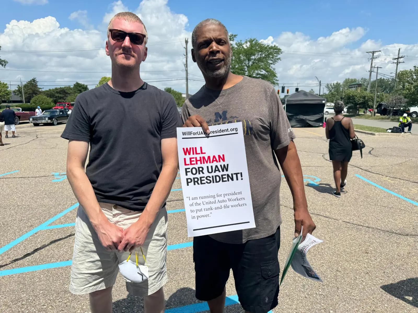 Will Lehman, in sunglasses and a dark T-shirt, stands with a supporter holding a “WILL LEHMAN FOR UAW PRESIDENT!” campaign sign in a parking lot, with vintage cars and event canopy tents in the background.