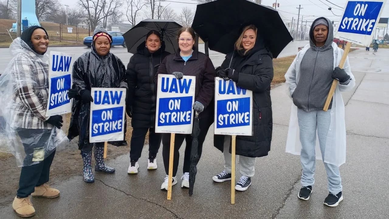 Six Lorain County JFS strikers holding blue and white “UAW On Strike” picket signs on a rainy roadside, several wearing rain ponchos and holding open umbrellas.
