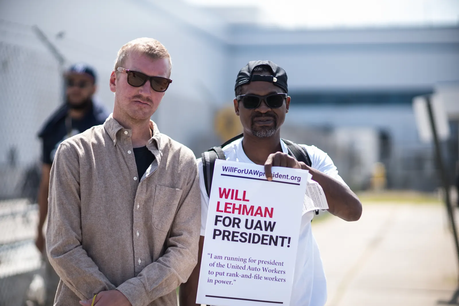 Will Lehman, in a tan shirt and sunglasses, stands outdoors near an industrial building beside a supporter in a backwards cap and sunglasses holding a sign that reads “WillForUAWpresident.org — WILL LEHMAN FOR UAW PRESIDENT! ‘I am running for president of the United Auto Workers to put rank-and-file workers in power.’”