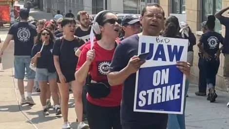 A picket line of Neighborhood Defender Service strikers on a city sidewalk; in front, a man holds a blue and white “UAW ON STRIKE” sign while a woman beside him in a red shirt chants.