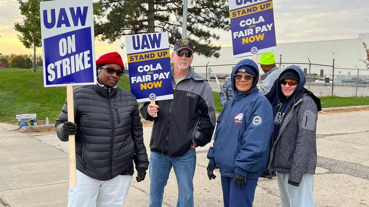 Four autoworkers in winter coats smiling on a picket line, holding “UAW ON STRIKE” and “STAND UP COLA FAIR PAY NOW” picket signs