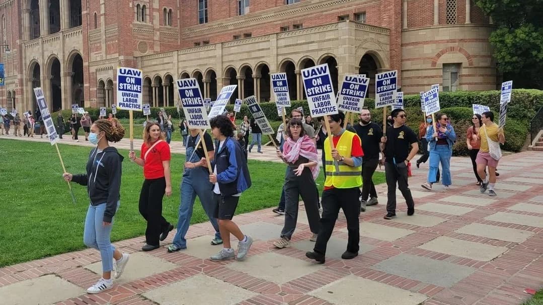 Academic workers picket outside a brick and stone university building, carrying blue and white signs reading “UAW ON STRIKE — UNFAIR LABOR PRACTICE.”