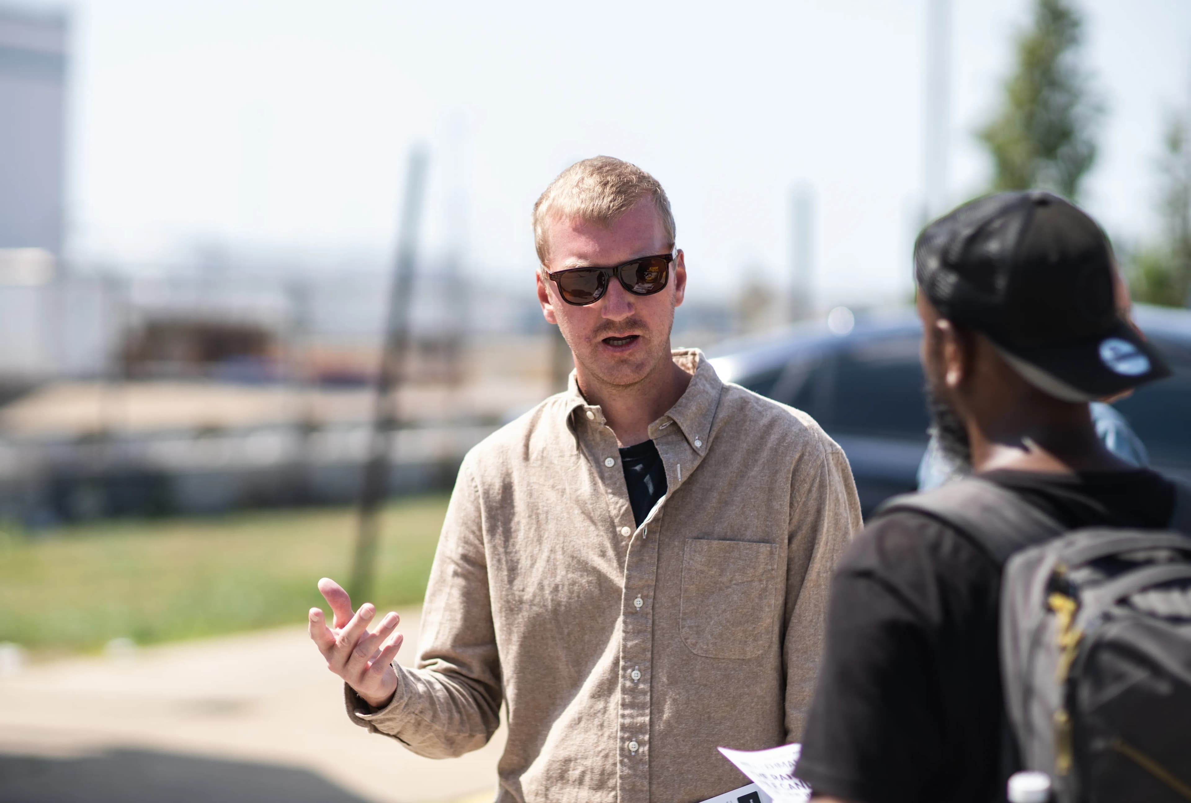 Will Lehman, in sunglasses and a tan button-down shirt, gestures while speaking with a worker wearing a backpack and a black cap in a parking lot outside a plant.