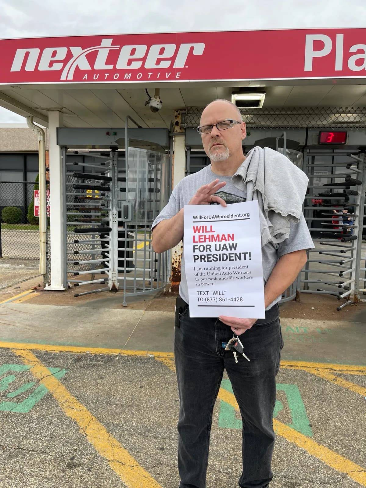 A worker stands outside the Nexteer Automotive plant entrance holding a 'Will Lehman for UAW President' flyer, with the Nexteer Automotive sign visible above the turnstile entrance behind him