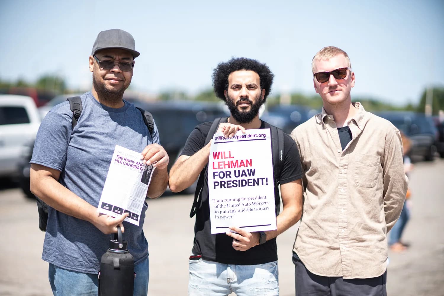 Will Lehman, right, stands with two autoworkers in a plant parking lot; the worker beside him holds a “WILL LEHMAN FOR UAW PRESIDENT!” poster reading “I am running for president of the United Auto Workers to put rank-and-file workers in power,” and another holds a campaign leaflet.