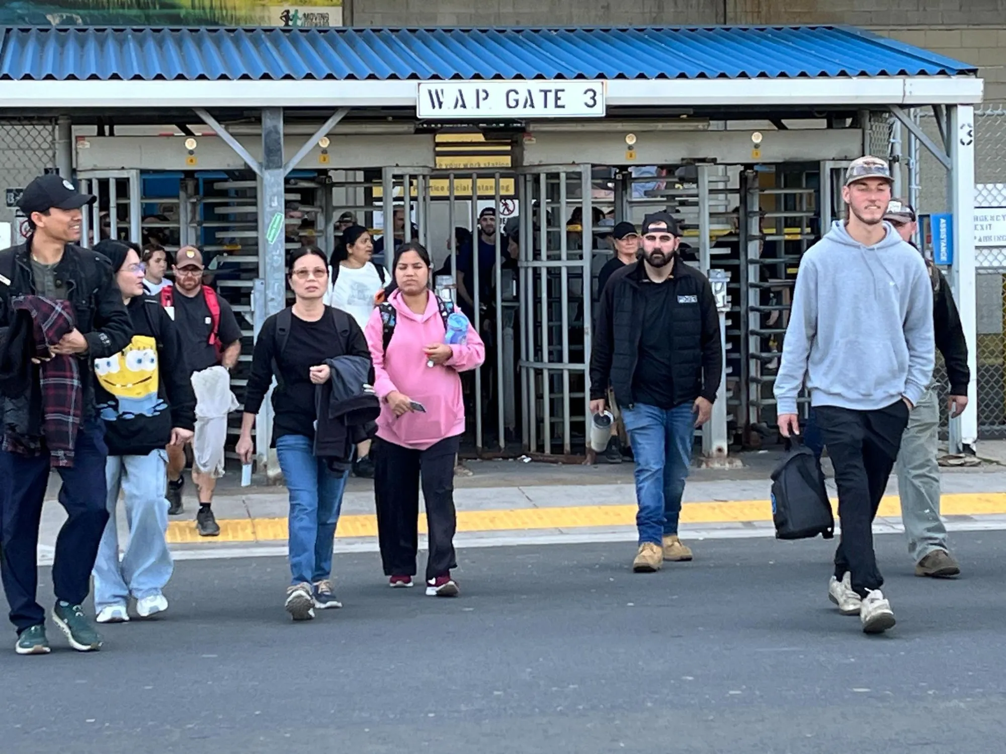Workers leaving the Windsor Assembly Plant through the turnstiles under a sign reading W.A.P. Gate 3, walking across the roadway toward the camera at shift change.