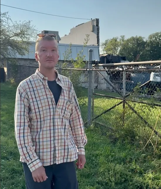 Will Lehman stands in a plaid shirt outside a chain-link fence at the Brent Industries plant in Toledo, with a debris-strewn lot and a building with a collapsed wall section visible behind the fence.