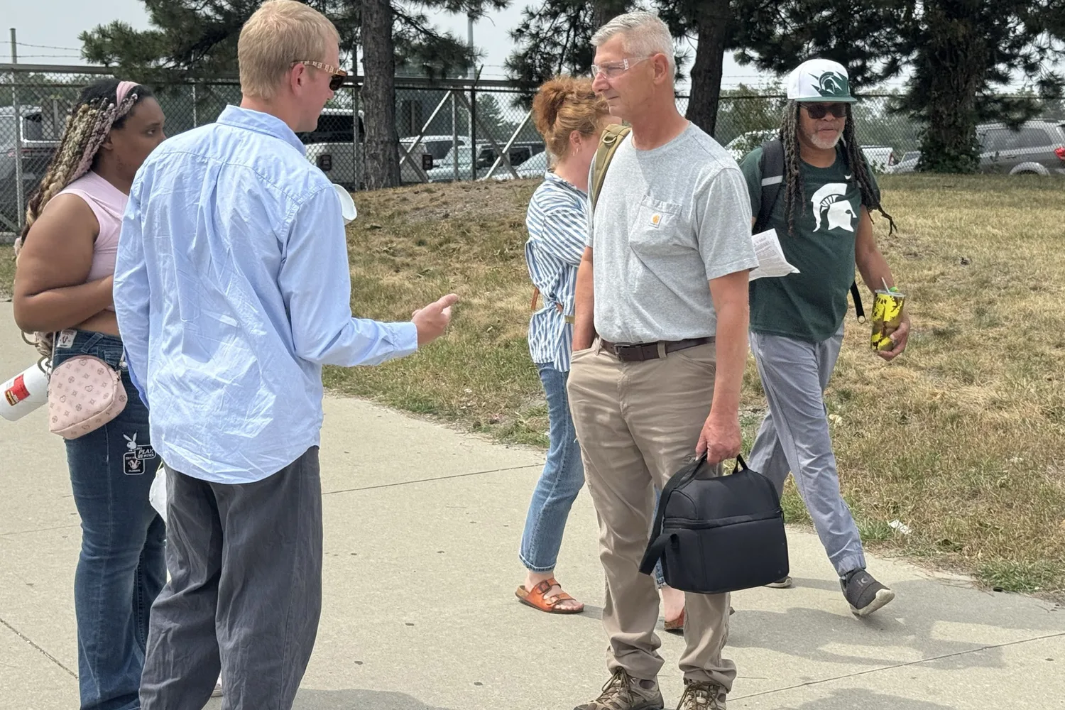 Will Lehman, in a light blue shirt and sunglasses, gestures as he talks with workers on a sidewalk outside the Stellantis Warren Truck Assembly Plant, while other workers walk past carrying campaign leaflets.