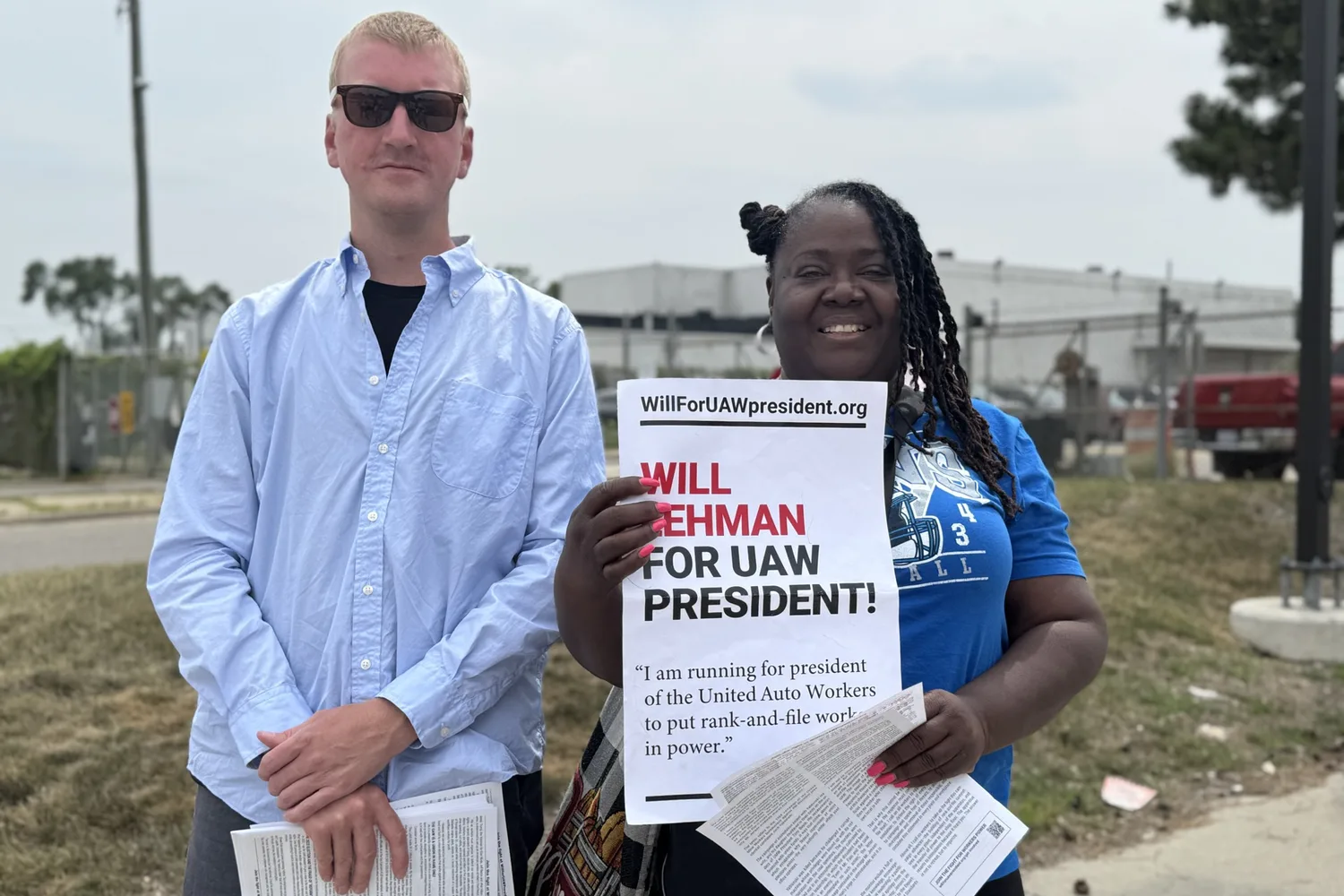 A worker in a blue T-shirt holds up a “WILL LEHMAN FOR UAW PRESIDENT!” poster and a handful of campaign leaflets while standing beside Will Lehman outside the Stellantis Warren Truck Assembly Plant.